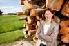 Landesrätin Michaela Langer-Weninger steht vor einem Holzstoß aus übereinandergeschichteten Baumstämmen, im Hintergrund Wiese, kleine Siedlung