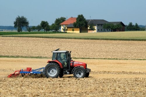 Im Vordergrund: Traktor eggt eine Ackerfläche, dahinter ein Bauernhof