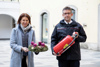 Feuerwehr-Landesrätin Langer Weninger und  Landes-Feuerwehrkommandant Robert Mayer stehen nebeneinander im Freien, Langer-Weninger hält einen Adventkranz in ihrem Händen, Robert Mayer einen Feuerlöscher, im Hintergrund Gebäude mit großen Bogenfenstern