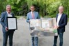 Karl Lehner, Landesrat Markus Achleitner und Alfred Mayr stehen nebeneinander auf einer asphaltierten Fläche und halten eine Urkunde bzw. ein großes Foto einer Gaststube mit Schank in Händen
