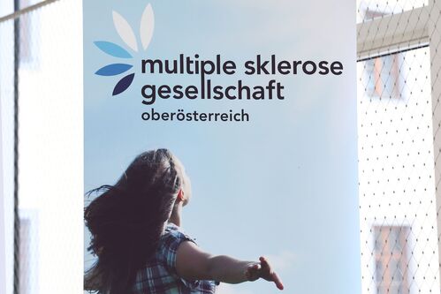 Schriftzug mit „multiple sklerose gesellschaft oberösterreich“ mit eine Frau von hinten, die ihre Arme ausbreitet