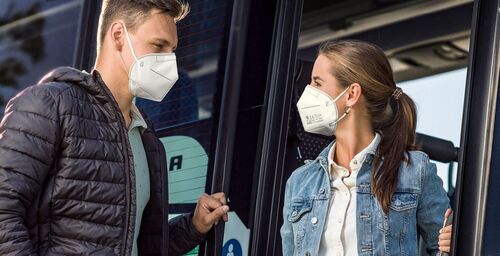 Junger Mann steht einer jungen Frau in einem öffentlichen Verkehrsmittel gegenüber; beide tragen eine FFP2-Maske
