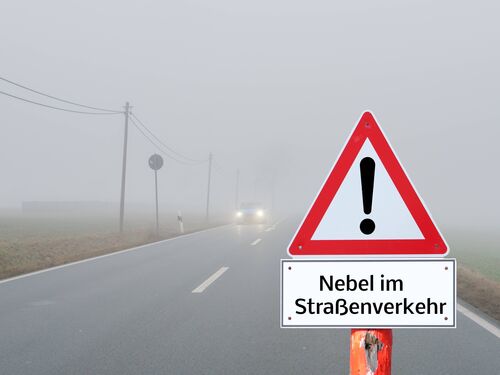Verkehrsschild „Achtung“ mit Zusatztafel „Nebel im Straßenverkehr“ an einer nebeligen Straße, auf der ein Auto mit eingeschalteten Scheinwerfern kommt