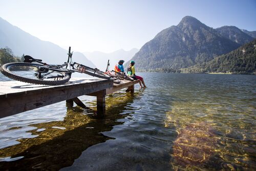 Eine Frau und ein Mann sitzen auf einem Steg und lassen die Füße in einen klaren Gebirgssee hängen, im Vordergrund liegen zwei Fahrräder auf dem Steg 