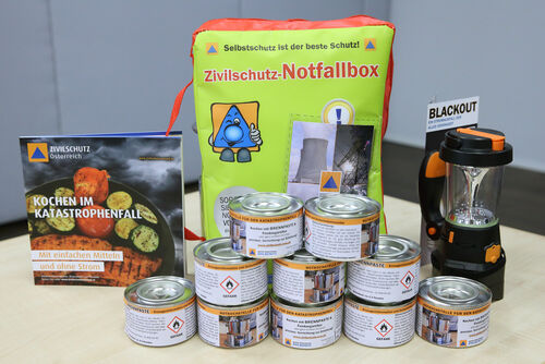 Auf einem Tisch steht ein Notfallradio, eine Broschüre „Blackout“,  eine Zivilschutz-Notfallbox, ein Buch „Kochen im Katastrophenfall“ und zehn Brennpasten für eine Notkochstelle.