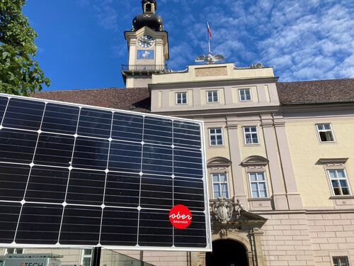 Photovoltaik-Panel steht auf einer Glasumrandung; darauf rechts unten befindet sich ein Logo des Land OÖ; im Hintergrund ist das Landhaus in Linz zu sehen.