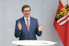 Wirtschafts- und Sport-Landesrat Markus Achleitner steht an einem Stehtisch und spricht mit erhobenen Händen – rechts neben ihm ist die Oberösterreich-Fahne zu sehen