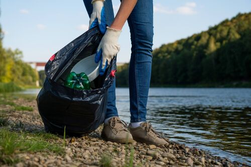 Frau sammelt an einem Flussufer Plastikmüll in einen Sack