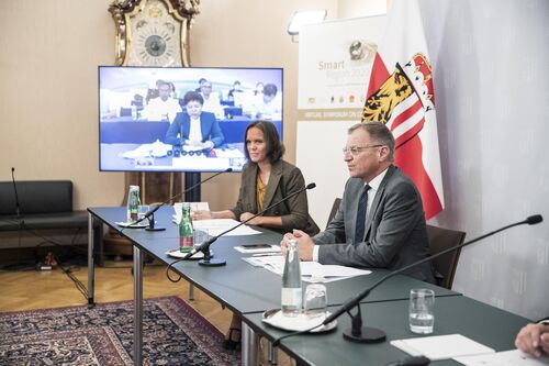 Moderatorin Nora Mack und LH Mag. Thomas Stelzer an einem Konferenztisch mit Mikrofonen, im Hintergrund Oberösterreich-Fahne und auf einem Bildschirm sichtbar die Live-Zuschaltung von REN Airong