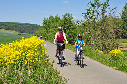 Frau und Kind fahren Rad auf einem Radweg in einer Landschaft mit Feldern, Wald, Hecken