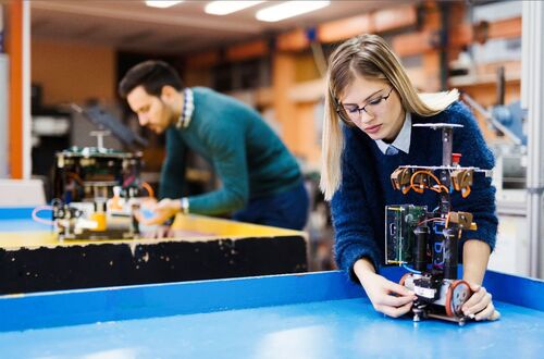 Eine Frau hält im Vordergrund in einem technischen Labor ein Computergerät auf einen Tisch. Dahinter steht an einem anderen Tisch ein Mann auch mit einem Computergerät.