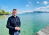 Landesrat Stefan Kaineder auf einem Steg an einem See, im Hintergrund Berge, Ortschaft, Kirche