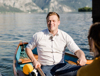 Landesrat Stefan Kaineder sitzt in einem Holzboot und hält jeweils ein Ruder in der Hand; im Hintergrund ist ein See im Salzkammergut zu sehen