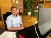 Landesrat Stefan Kaineder sitzt an einem Schreibtisch vor einem iPad, drahtlose Kopfhörer im Ohr, im Hintergrund Regal mit Zimmerpflanze und Pinnwand mit Bildern.