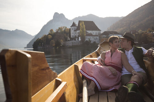 Ein Paar in Tracht liegt in einer Zille auf dem Traunsee, im Hintergrund eine Kirche, herbstliche Wälder, Berge