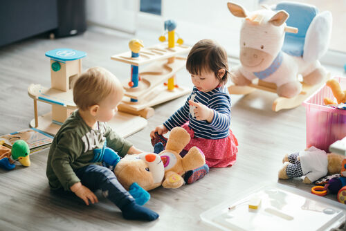 Zwei kleine Kinder sitzen am Boden und spielen mit einem Plüschtier; neben ihnen sind verschiedene Spielsachen zu sehen. 