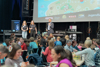 Landesrat Dr. Wolfgang Hattmannsdorfer steht auf einer Bühne und spricht in ein Mikrofon, neben ihm zwei weitere Personen, im Vordergrund Publikum spielend an Tischen