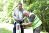 Landesrat Mag. Günther Steinkellner mit Wildwarngerät und Hannes Langmayr mit Akkuschrauber stehen am Straßenrand bei einem Leitpflock, im Hintergrund Wald