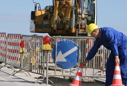 Bauarbeiter stellt an einer Straßenbaustelle Warnzylinder auf, Absperrgitter, Bagger