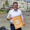 Landesrat Stefan Kaineder steht auf dem Linzer Hauptplatz und hält ein kleines Poster mit Beschriftung Viel Falt in Concert in Händen, im Hintergrund Häuser, Pestsäule, Menschen, Blumenbeete