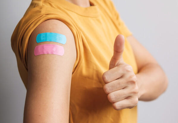 Nahaufnahme eines Oberarms mit zwei bunten Pflastern an der Impfstelle; die Person trägt ein gelbes T-Shirt. Daneben zeigt eine Hand den Daumen nach oben