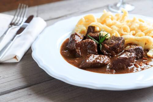 Ein Teller Wildfleischgulasch mit Nockerl auf einem Tisch, daneben liegen eine Serviette, Gabel und Messer.