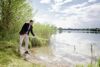 Landesrat Stefan Kaineder steht an einem Seeufer und taucht ein Gefäß, das an einer langen Stange befestigt ist, ins Wasser