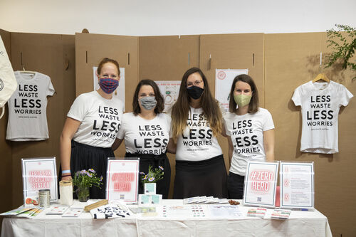 Vier Frauen stehen nebeneinander vor einem Stand und tragen T-Shirts mit der Aufschrift „Less Waste Less Worries“