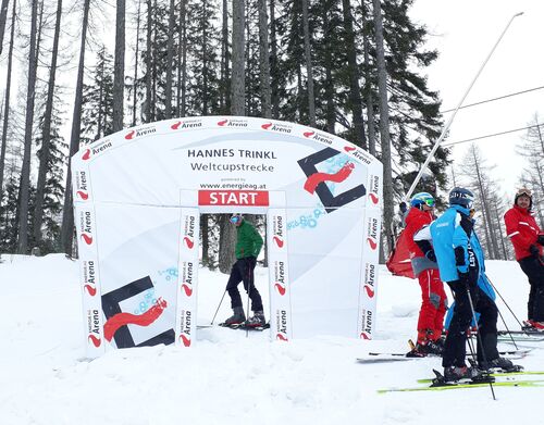 Starthaus mit der Aufschrift „Hannes Trinkl Weltcupstrecke“ flankiert von mehreren Skifahrern.
