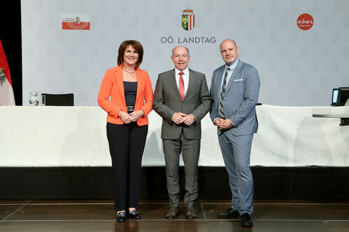 Sabine Binder, Max Hiegelsberger und Peter Binder stehen nebeneinander vor einem Konferenztisch, im Hintergrund Fotowand mit dem Logo für den Oö. Landtag, Oberösterreich-Wappen und Oberösterreich-Symbol