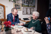 Landeshauptmann Mag. Thomas Stelzer sitzt neben Theresia Stadlmayr an einem gedeckten Tisch in ihrem Wohnzimmer und gratuliert zu ihrem 105. Geburtstag. An der Wand hängen Bilder.
