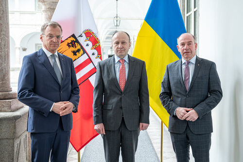 Landeshauptmann Mag. Thomas Stelzer, Botschafter Dr. Vasyl Khymynets und Landtagspräsident Max Hiegelsberger im Gang des Arkadenhofs des Landhauses in Linz, im Hintergrund die oberösterreichische und die ukrainische Fahne.