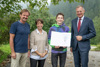 Ein Mann und eine Frau, Simon Lehner-Ölweiner mit Urkunde und Landeshauptmann Mag. Thomas Stelzer stehen nebeneinander auf einem geschotterten Bereich, im Hintergrund Wald, Bäume