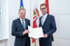 Landeshauptmann Mag. Thomas Stelzer und Dr. Florian Kolmhofer stehen vor einer EU-, Oberösterreich- und Österreich-Fahne und halten gemeinsam die Bestellungsurkunde in Händen