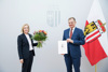 Mag.a. Carmen Breitwieser und Landeshautmann Mag. Thomas Stelzer mit Blumen und Dekret in Händen, im Hintergrund Oberösterreich-Fahne