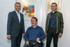Landesrat Wolfgang Hattmannsdorfer, Andreas Anderle im Rollstuhl und Gerald Rechberger stehen nebeneinander vor einer Wand, an der zwei bunte Bilder hängen