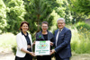 Landesrätin Michaela Langer-Weninger Stadträtin Eva Schobesberger und Bürgermeister Klaus Luger stehen nebeneinander vor einem kleineren Nadelbaum und halten gemeinsamein Schild mit Beschriftung Natur Garten Oberösterreich, Natur im Garten Gemeinde