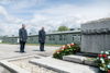 Landeshauptmann Mag. Thomas Stelzer und Bundespräsident Dr. Alexander Van der Bellen stehen mit gesenktem Kopf vor einem Denkmal, vor dem ein großer Kranz liegt, im Hintergrund Baracken-Gebäude