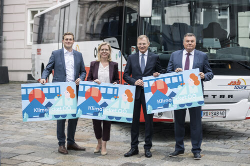 Landesrat Stefan Kaineder, Ministerin Leonore Gewessler, Landeshauptmann Mag. Thomas Stelzer und Landesrat Mag. Günther Steinkellner stehen nebeneinander auf einem Platz vor dem Landhaus, hinter ihnen ein großer Linienbus, gemeinsam halten sie Schilder mit Beschriftung KlimaTicket OÖ