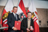 Landeshauptmann Mag. Thomas Stelzer und Franz Josef Altenburg stehen nebeneinander mit Dokument und Medaille in Händen, im Hintergrund zwei Oberösterreich- und eine Österreichfahne