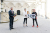 LH Mag. Stelzer mit einem Blumenstrauß, Mama Elisabeth und ihr Sohn Timo halten ein Plakat mit einer Ankündigung für die OÖ. Landesausstellung hoch, im Hintergrund Brunnen und Arkadenhof des Landhauses.