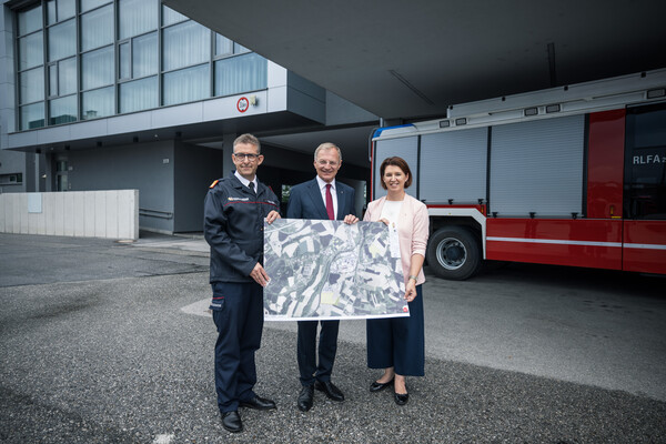 Landes-Feuerwehrkommandant Robert Mayer, Landeshauptmann Thomas Stelzer und Feuerwehr-Landesrätin Michaela Langer-Weninger halten gemeinsam den Lageplan des zukünftigen Landes-Feuerwehrzentrums; im Hintergrund ist ein rotes Feuerwehrfahrzeug zu sehen.