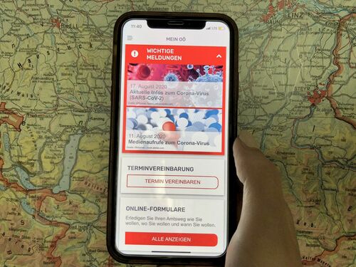 Hand hält ein Mobiltelefon, auf dem die Startseite der App sichtbar ist, im Hintergrund Ausschnitt einer Oberösterreich-Karte