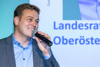 Landesrat Stefan Kaineder steht vor einer Videowand mit Beschriftung Landesrat Stefan Kaineder Oberösterreich und spricht in ein Mikrofon