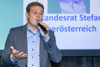 Landesrat Stefan Kaineder steht vor einer Videowand mit Beschriftung Landesrat Stefan Kaineder Oberösterreich und spricht in ein Mikrofon