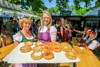 Viele Menschen auf einem Grünmarkt mit Verkaufsständen, im Vordergrund zwei Frauen mit einem Tablett voller Bauernkrapfen