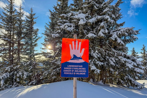 Rot-blaues Warnschild mit der Aufschrift „Lawinengefahr“ vor einer Wald- und Schneelandschaft.