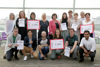 Gruppenfoto, 16 Menschen in zwei Reihen, Landesrätin Birgit Gerstorfer gemeinsam mit allen Preisträgerinnen und Preisträgern vor einer Glasfront mit Blick auf Häuser und Fluss