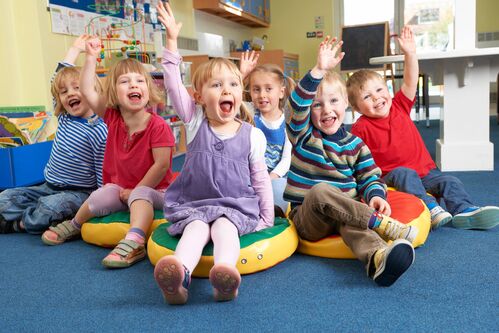 Sechs Kinder sitzen auf Polstern in einem Kindergartenraum und zeigen begeistert auf