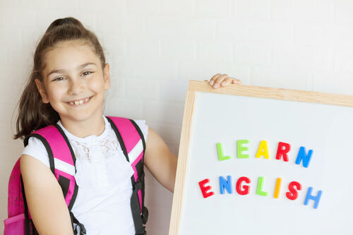 Schülerin mit umgehängter Schultasche hält eine Tafel mit der Aufschrift „Learn English“ in den Händen